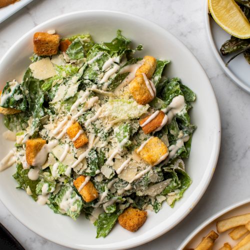 A Caesar salad with croutons and dressing on a plate, accompanied by a side of fries and lemon wedges on a marble table.