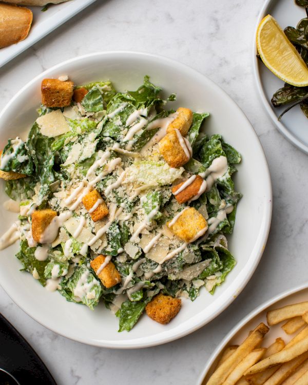 A Caesar salad with croutons and dressing on a plate, accompanied by a side of fries and lemon wedges on a marble table.