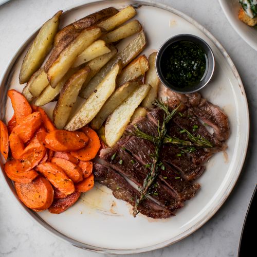 A plate with sliced steak, roasted potato wedges, and carrots, accompanied by a small dish of sauce on the side.