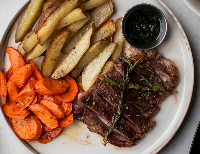 A plate with sliced steak, roasted potato wedges, and carrots, accompanied by a small dish of sauce on the side.