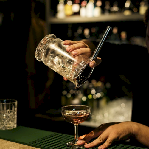 A bartender is pouring a drink from a mixing glass into a cocktail glass on a bar counter with shelves of bottles in the background.