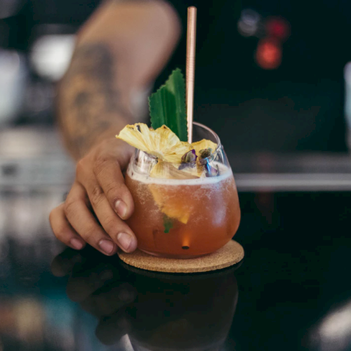 A person is serving a cocktail with a pineapple garnish in a glass, resting on a cork coaster with a straw.