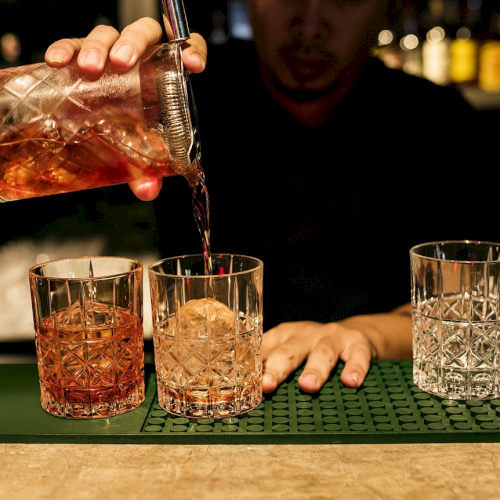 A bartender is pouring drinks from a mixing glass into three glasses with ice on a bar counter.