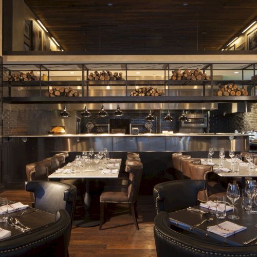 A warmly lit, modern restaurant interior with elegant table settings, wooden accents, and an open kitchen.