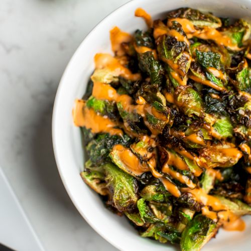 A bowl of roasted Brussels sprouts drizzled with a creamy orange sauce is placed on a marble surface.