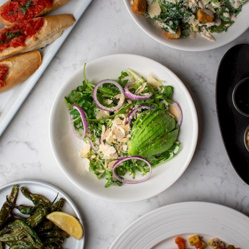 An assortment of dishes: salad with avocado, Caesar salad, roasted peppers with lemon, bruschetta, and a plate with rice and vegetables.