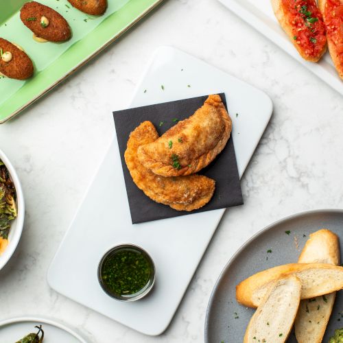 A spread of tapas: empanadas, croquetas, bread with tomato, toasted bread, peppers, and a green dipping sauce on white plates and bowls.