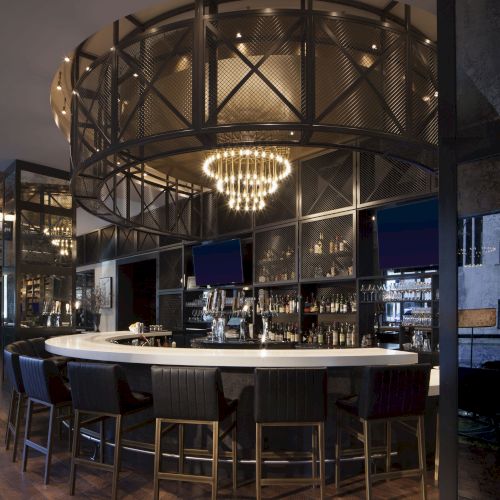 The image shows a stylish bar with a round counter, high chairs, and a chandelier overhead, creating an inviting atmosphere.