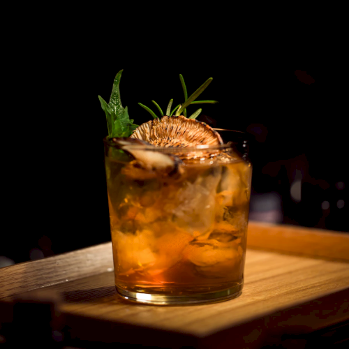 A cocktail glass with ice, garnished with herbs and a dried citrus slice, sits on a wooden tray against a dark background.