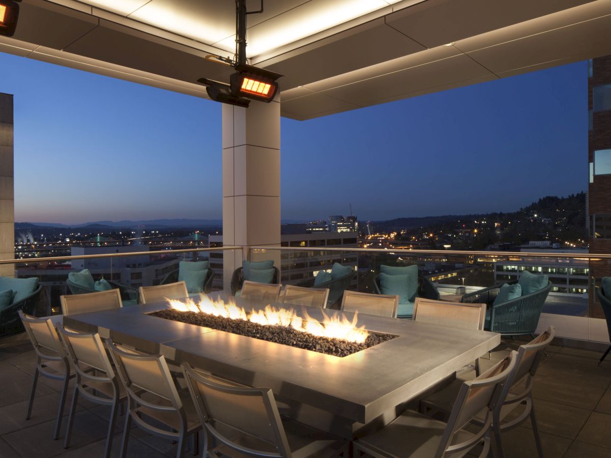 Rooftop patio with a long table featuring a fire feature, surrounded by chairs, with a cityscape view at dusk.