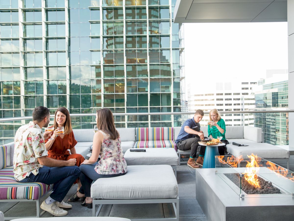 People are socializing on a rooftop patio with a modern fire pit, seating, and urban buildings in the background.