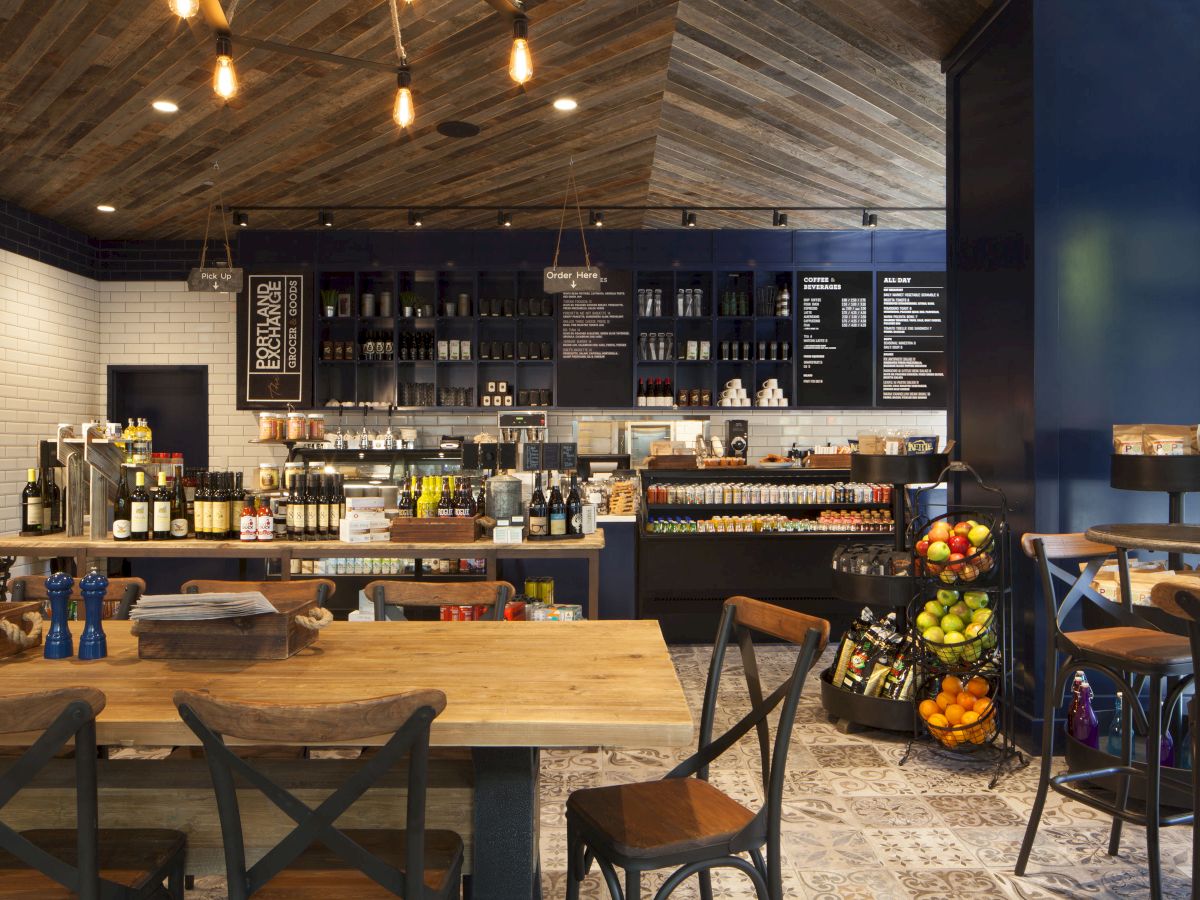 The image shows a cozy café interior with wooden tables, shelves of products, a fruit stand, and a menu board on the wall.