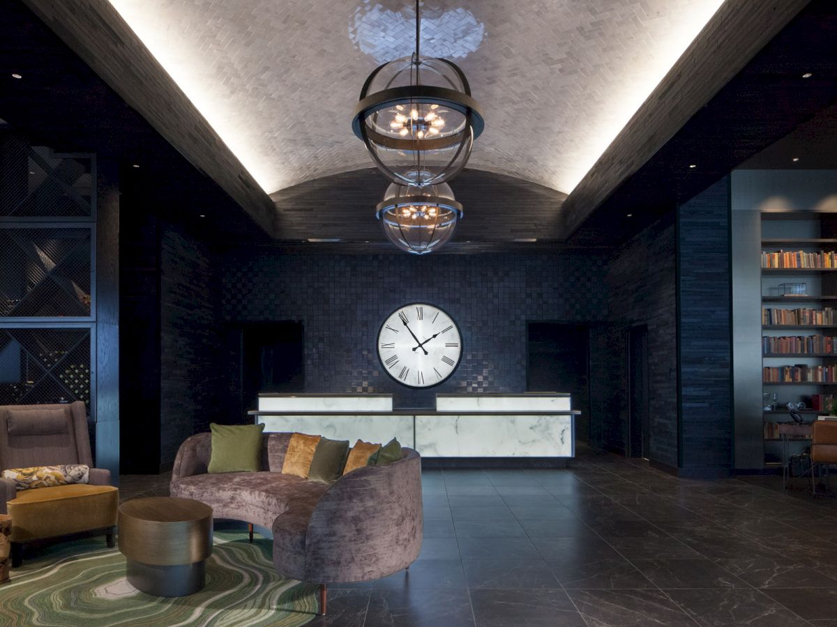 A stylish hotel lobby with modern seating, a large wall clock, and bookshelves, illuminated by chic ceiling lights.